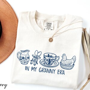 A white t-shirt with a graphic design of various kitchen utensils and a chicken, along with the text "IN MY GRANNY ERA" printed on it. The t-shirt is placed on a white surface, and there is a brown hat and a plant nearby.
