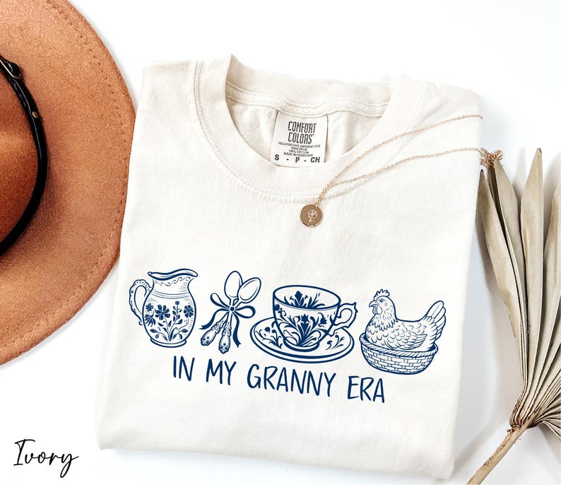 A white t-shirt with a graphic design of various kitchen utensils and a chicken, along with the text "IN MY GRANNY ERA" printed on it. The t-shirt is placed on a white surface, and there is a brown hat and a plant nearby.
