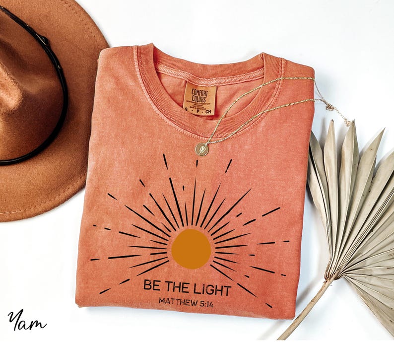 K&ouml;nnte beinhalten: Ein korallenfarbenes T-Shirt mit einer schwarzen Sonnengrafik und dem Text "Sei das Licht Matth&auml;us 5:16".