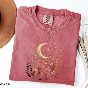 a pink t-shirt with a crescent moon and starry night sky design, along with some dried flowers and leaves. The t-shirt is placed on a white surface, and there is a brown hat and a plant in the background.