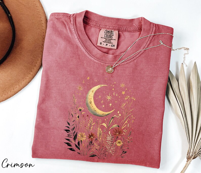 a pink t-shirt with a crescent moon and starry night sky design, along with some dried flowers and leaves. The t-shirt is placed on a white surface, and there is a brown hat and a plant in the background.