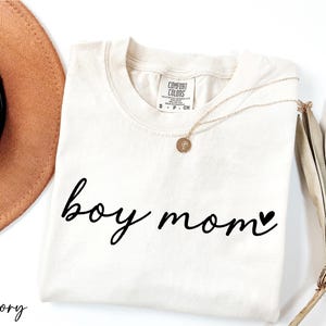 A white t-shirt with the text "boy mom" printed on it, along with a necklace and a hat, all placed on a white background.