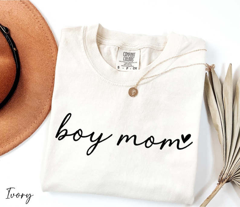 A white t-shirt with the text "boy mom" printed on it, along with a necklace and a hat, all placed on a white background.