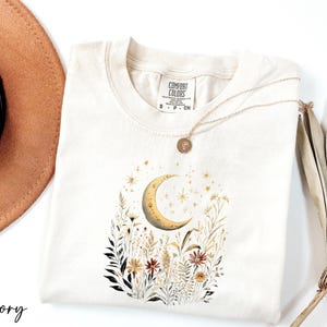 a white t-shirt with a floral design and a crescent moon, along with a brown hat and a fan.