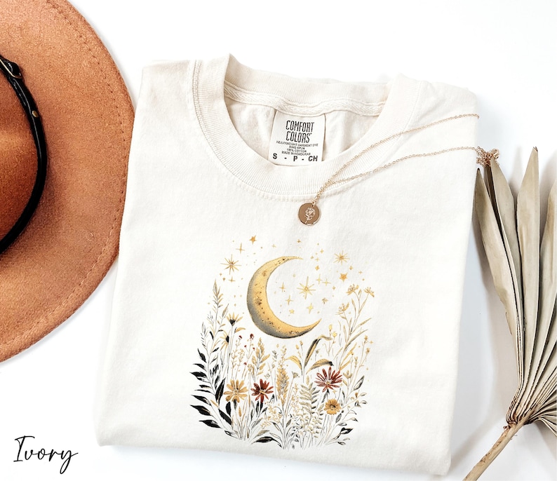a white t-shirt with a floral design and a crescent moon, along with a brown hat and a fan.
