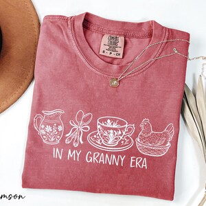 A red t-shirt with white text and illustrations, including a teapot, spoon, cup, and chicken, along with the text "IN MY GRANNY ERA". The t-shirt is placed on a white surface, with a brown hat and some dried leaves nearby.