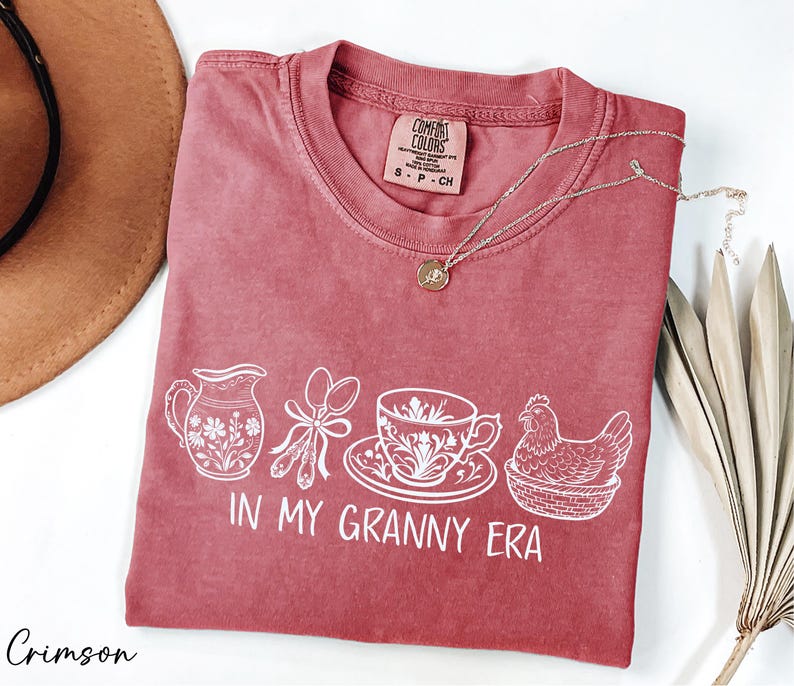 A red t-shirt with white text and illustrations, including a teapot, spoon, cup, and chicken, along with the text "IN MY GRANNY ERA". The t-shirt is placed on a white surface, with a brown hat and some dried leaves nearby.