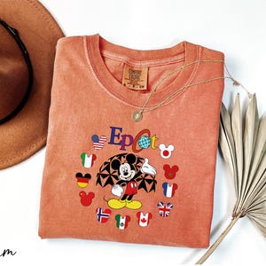 May include: A coral orange t-shirt with a graphic of Mickey Mouse in front of the Epcot World Showcase. Mickey Mouse is wearing a red and yellow outfit and is surrounded by flags representing different countries. The text "Epcot" is printed in black above Mickey Mouse.