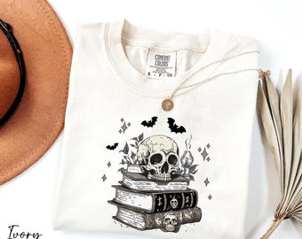 May include: Ivory-colored Comfort Colors t-shirt featuring a graphic of a skull atop a stack of books, with bats and stars. A gold necklace and a dried plant are also visible. The word "Ivory" is printed at the bottom.