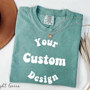 Op de afbeelding: Lichtgroen T-shirt met de tekst "Your Custom Design" in wit, retro-lettertype. Het shirt is gemaakt van een zacht, comfortabel materiaal.