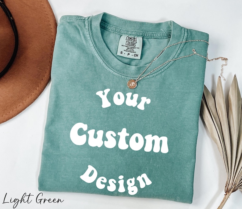 Op de afbeelding: Lichtgroen T-shirt met de tekst "Your Custom Design" in wit, retro-lettertype. Het shirt is gemaakt van een zacht, comfortabel materiaal.