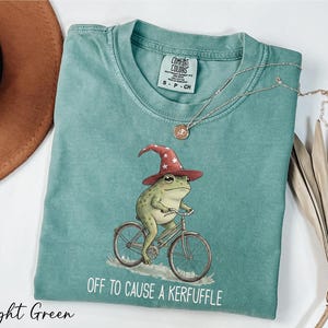 A light green t-shirt with a whimsical illustration of a frog riding a bicycle, wearing a wizard's hat, and the text "Off to cause a Keruffle" printed on it.