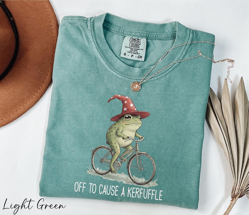 A light green t-shirt with a whimsical illustration of a frog riding a bicycle, wearing a wizard's hat, and the text "Off to cause a Keruffle" printed on it.