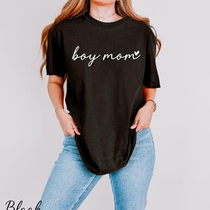 A woman wearing a black t-shirt with the text "boy mom" printed on it, standing against a plain background.