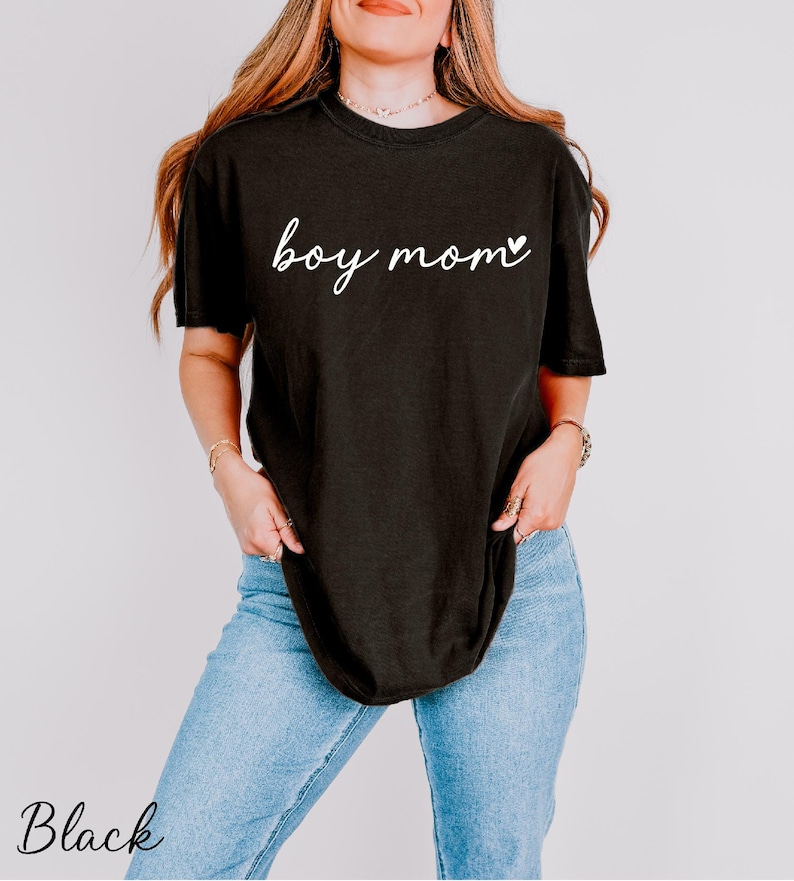 A woman wearing a black t-shirt with the text "boy mom" printed on it, standing against a plain background.