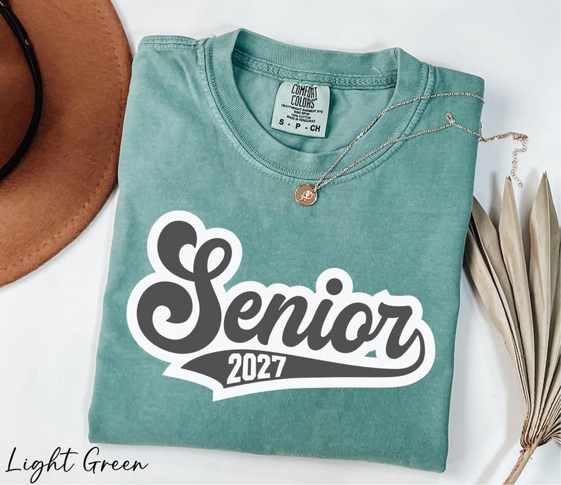 A light green t-shirt with a black and white text design that says "Senior 2027" is displayed on a white background.