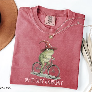 a red t-shirt with a whimsical illustration of a frog riding a bicycle, accompanied by the text "Off to cause a Keruffle".