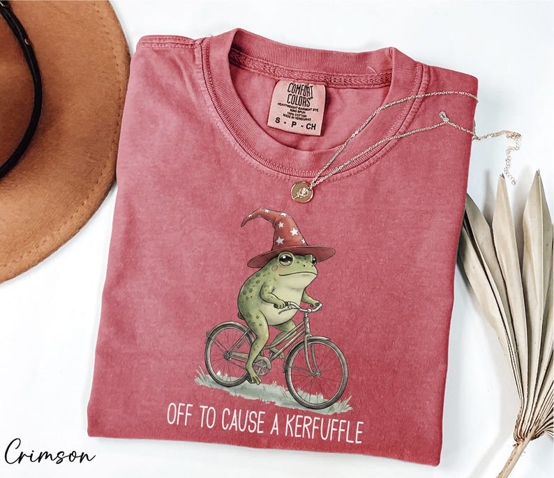a red t-shirt with a whimsical illustration of a frog riding a bicycle, accompanied by the text "Off to cause a Keruffle".