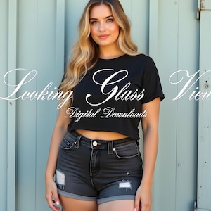 Puede incluir: Una mujer con una camiseta negra recortada con el texto "Looking Glass Views Digital Downloads" y pantalones cortos de mezclilla oscura con detalles desgastados. Está de pie frente a una pared azul claro.