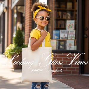 以下が含まれることがあります： サングラスと黄色のシャツを着た若い人が、クリーム色のトートバッグを持っています。バッグには「Looking Glass Views」と「Digital Downloads」の文字がプリントされています。背景にはレンガ造りの建物と本屋があります。