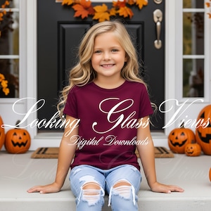 Può includere: Una ragazza indossa una maglietta bordeaux con la scritta "Looking Glass Views Digital Downloads" in corsivo bianco. È seduta su un gradino del portico, con zucche e foglie autunnali sullo sfondo. Indossa jeans blu strappati.