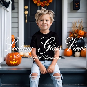 May include: A young child wearing a black t-shirt and ripped blue jeans sits on a porch. The shirt has the text "Looking Glass Views" in white script. A carved pumpkin and other pumpkins are nearby, suggesting a Halloween theme.