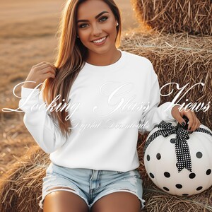May include: A person wearing a white sweatshirt and denim shorts sits in front of a hay bale. They hold a white pumpkin decorated with black polka dots and a black and white ribbon. The text "Looking Glass Views Digital Downloads" is on the sweatshirt.
