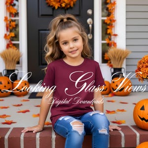 May include: A young girl in a maroon t-shirt and ripped blue jeans sits on a brick step. The shirt features the words "Looking Glass Views" and "Digital Downloads" in white script. Pumpkins and autumn leaves decorate the porch.