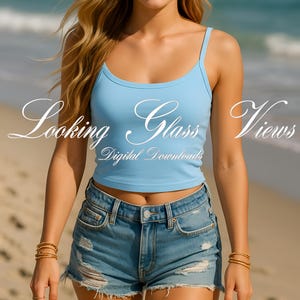 May include: A woman wearing a light blue tank top and denim shorts. The tank top is cropped and has a thin strap. The shorts are distressed and have a frayed hem. The woman is standing on a sandy beach with the sea in the background. The text "Looking Glass Views" is superimposed on the image.