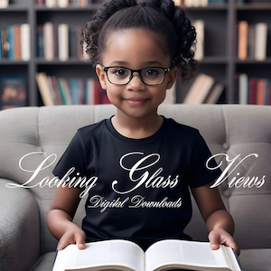 May include: A young girl with dark brown hair wearing black framed glasses sits on a grey sofa and reads a book. The book is open and the pages are white. The girl is wearing a black t-shirt with the text "Looking Glass Views Digital Downloads" on it.