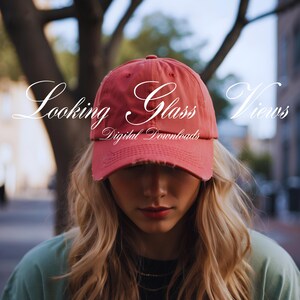 May include: A woman wearing a red baseball cap with the text "Looking Glass Views Digital Downloads" on it. She has long blonde hair and is looking down.