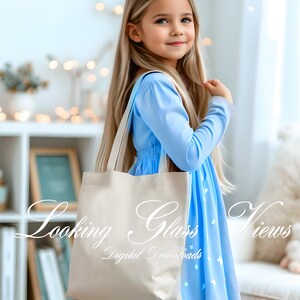 Op de afbeelding: Een jong meisje in een lichtblauwe jurk met witte stippen houdt een beige tote bag vast. De tas heeft de tekst "Looking Glass Views" en "Digital Downloads" erop gedrukt.