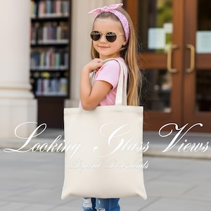 以下が含まれることがあります： 「Looking Glass Views Digital Downloads」の文字が筆記体で書かれたクリーム色のトートバッグ。バッグには長い持ち手が付いています。画像には、サングラスとピンクのヘアバンドを身に着けた若い人がトートバッグを持っている様子が写っています。