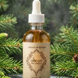 Può includere: Una bottiglia di vetro trasparente di amari di punta di abete con un contagocce bianco. L'etichetta recita "Ocean Mist Infusions" e "Klawock, AK". La bottiglia è riempita con un liquido marrone dorato ed è circondata da rami di pino verdi.