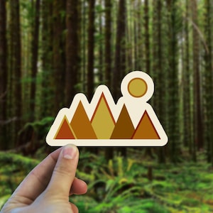 May include: A sticker with a stylized mountain range design in shades of brown, orange, and yellow. A yellow sun is above the mountains.