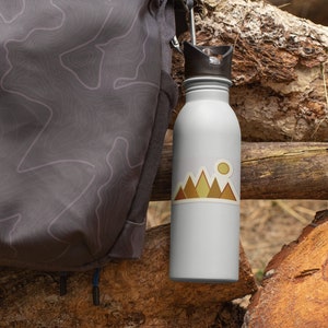 May include: A white water bottle with a brown mountain and sun sticker. The bottle is attached to a gray backpack with a topographic map design.