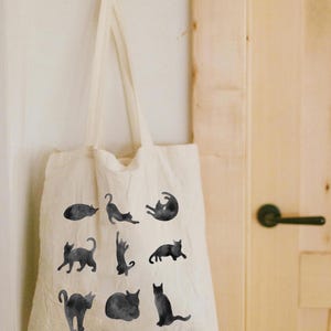 May include: A cream-colored tote bag with black watercolor illustrations of cats in various poses. The bag is hanging on a white wall. The cats are in different positions, such as lying down, stretching, and sitting.