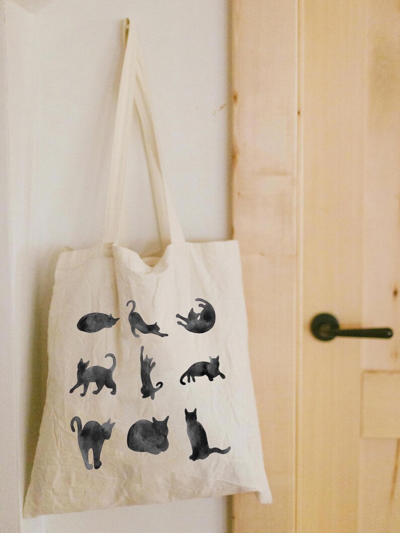 May include: A cream-colored tote bag with black watercolor illustrations of cats in various poses. The bag is hanging on a white wall. The cats are in different positions, such as lying down, stretching, and sitting.
