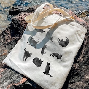 May include: A cream-colored tote bag featuring black cat silhouettes in various poses. The bag has two cream-colored handles and is resting on a large rock near water. The cats are in different positions, some standing, some lying down.