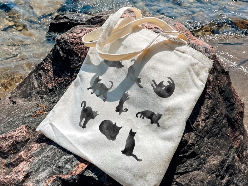 May include: A cream-colored tote bag featuring black cat silhouettes in various poses. The bag has two cream-colored handles and is resting on a large rock near water. The cats are in different positions, some standing, some lying down.