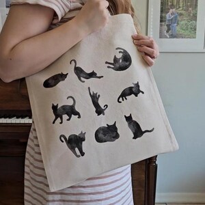 May include: A beige tote bag featuring black cat illustrations in various poses. The cats are depicted in a watercolor style, adding a whimsical touch to the bag. The bag is made of a sturdy fabric, suitable for carrying groceries or personal items.
