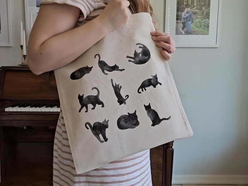 May include: A beige tote bag featuring black cat illustrations in various poses. The cats are depicted in a watercolor style, adding a whimsical touch to the bag. The bag is made of a sturdy fabric, suitable for carrying groceries or personal items.