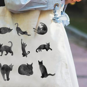 May include: A cream-colored tote bag featuring a repeating pattern of black cat silhouettes in various poses. The cats are depicted in a watercolor style, adding a soft, artistic touch to the design. The bag is held by a person wearing a light blue shirt.