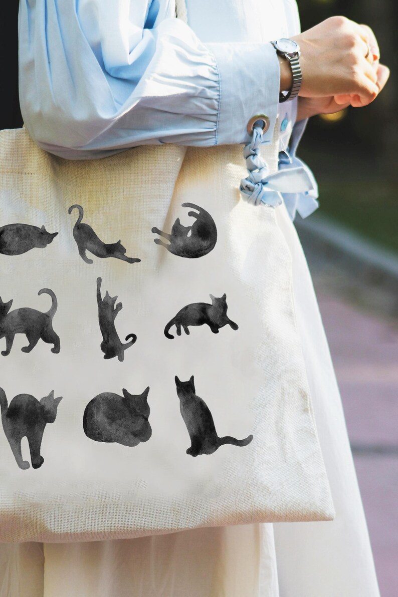 May include: A cream-colored tote bag featuring a repeating pattern of black cat silhouettes in various poses. The cats are depicted in a watercolor style, adding a soft, artistic touch to the design. The bag is held by a person wearing a light blue shirt.