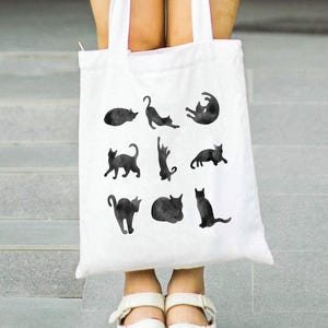 May include: White tote bag featuring black watercolor illustrations of cats in various poses. The bag has a simple design with a rectangular shape and two handles. The cats are depicted in different positions, such as stretching, sitting, and curled up.