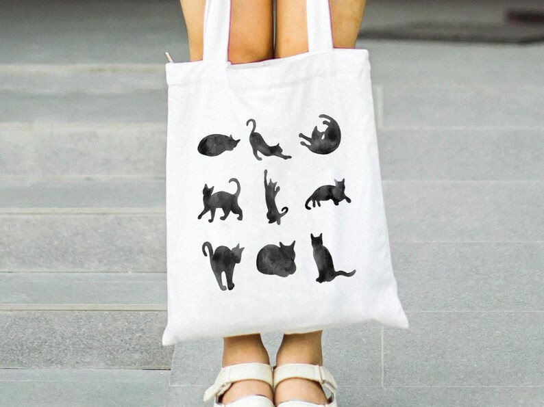 May include: White tote bag featuring black watercolor illustrations of cats in various poses. The bag has a simple design with a rectangular shape and two handles. The cats are depicted in different positions, such as stretching, sitting, and curled up.
