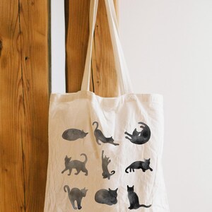 May include: Cream-colored tote bag featuring black watercolor cat illustrations in various poses. The bag is hanging against a wooden background. The bag has two long handles.