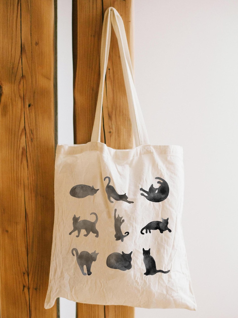 May include: Cream-colored tote bag featuring black watercolor cat illustrations in various poses. The bag is hanging against a wooden background. The bag has two long handles.