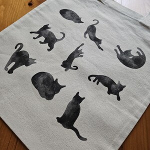 May include: A beige tote bag featuring black cat silhouettes in various poses. The cats are printed across the front of the bag, which has two beige handles. The bag is made of a sturdy fabric and is suitable for carrying groceries or other items.