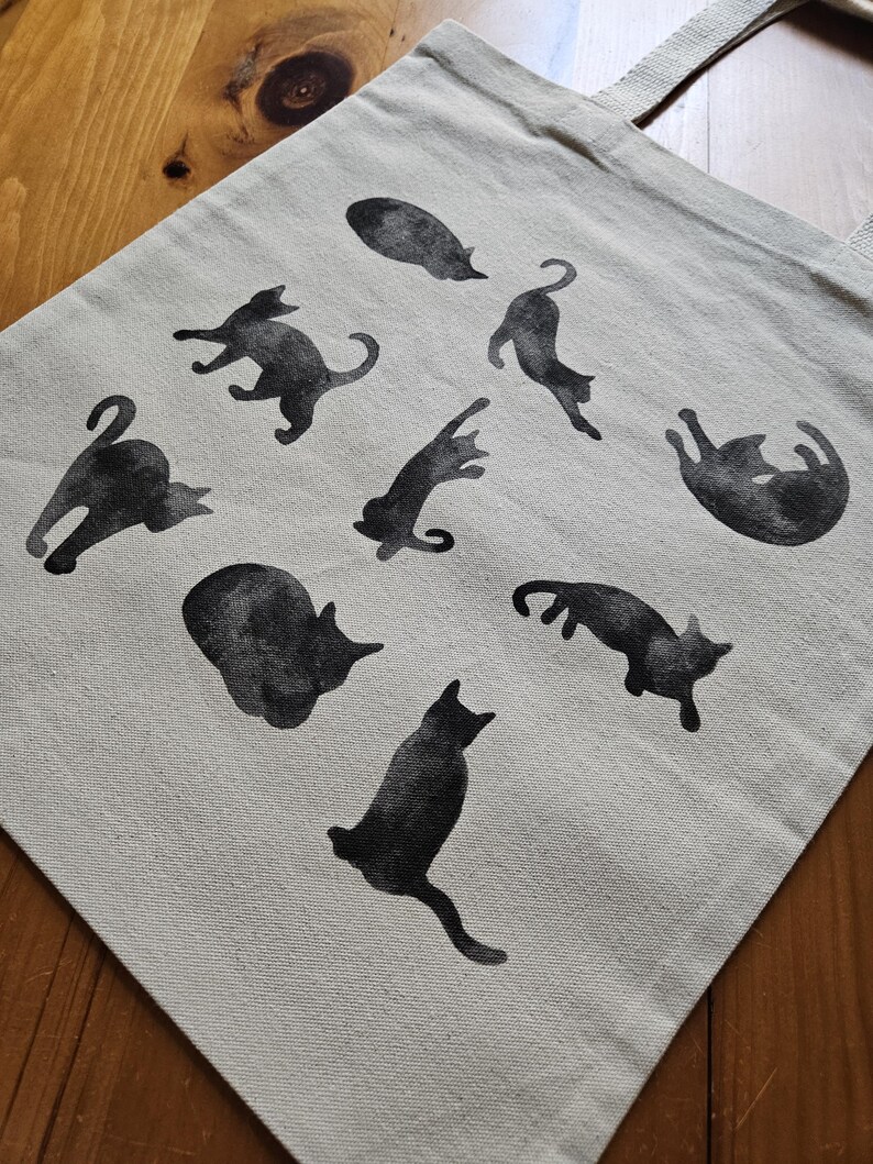 May include: A beige tote bag featuring black cat silhouettes in various poses. The cats are printed across the front of the bag, which has two beige handles. The bag is made of a sturdy fabric and is suitable for carrying groceries or other items.
